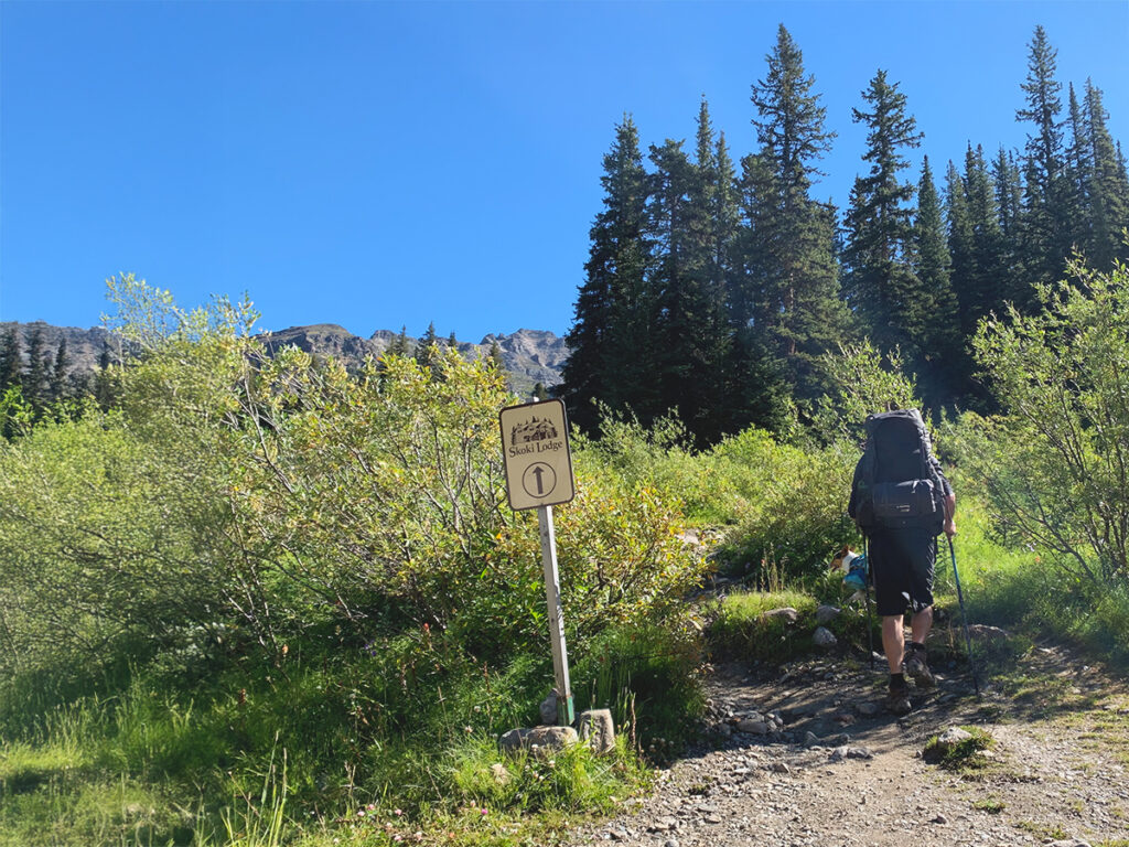 A man wearing a large hiking backpack hikes up a trail past a distance marker sign